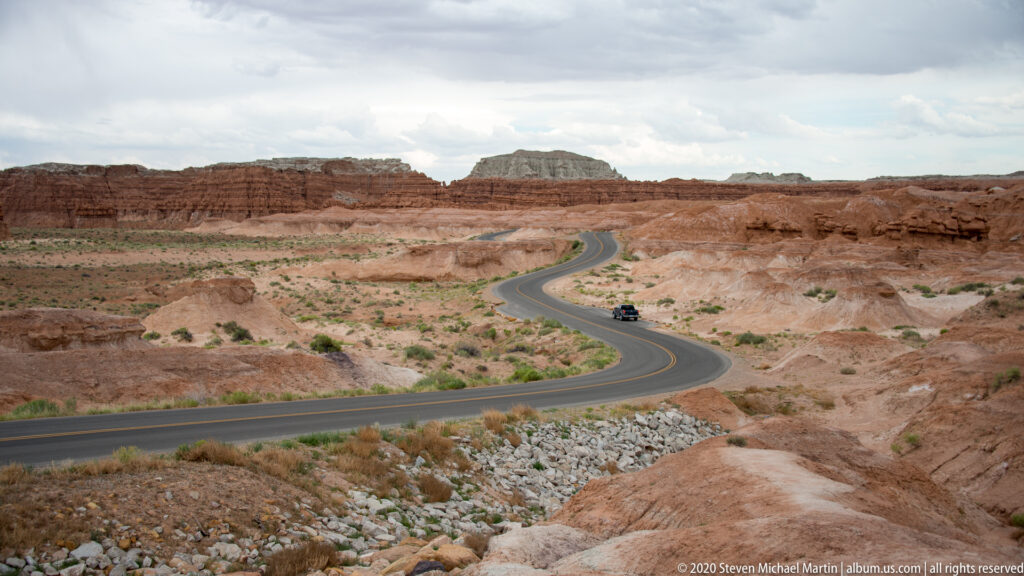 A winding paved road crosses pale badlands toward distant mesas, with a vehicle on the curve.