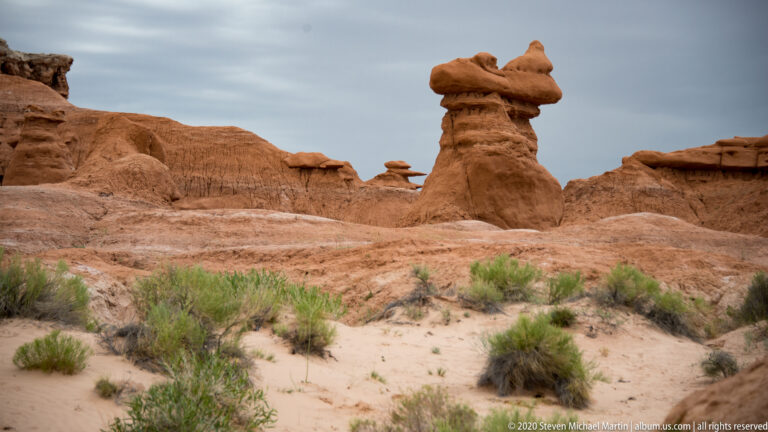 A tall irregular hoodoo rises among smaller sandstone forms and low shrubs in a sandy basin.