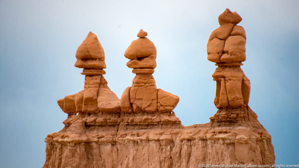 Three narrow capped hoodoos rise from a sandstone escarpment above a low ridge of rounded rocks.