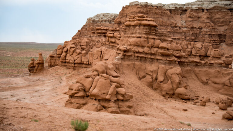 Rounded hoodoos cluster below a tall banded cliff, with a small shrub in the foreground.