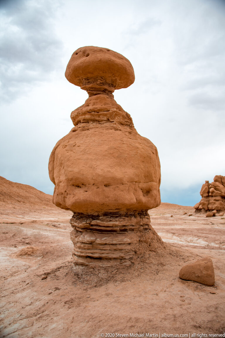 A wide rounded capstone sits atop a layered pedestal on an open sandstone plain.