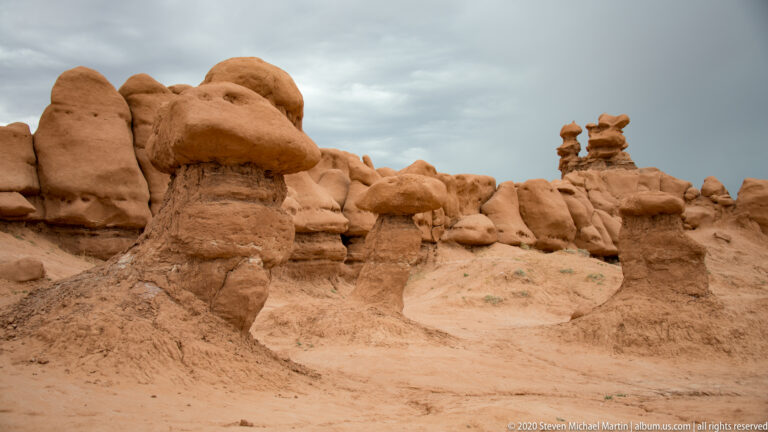 Rounded sandstone hoodoos of different heights stand across a sandy basin beneath a pale overcast sky.