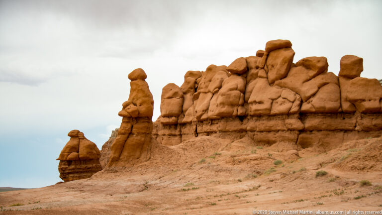 A ridge of blocky sandstone caps rises above two isolated hoodoos under a narrow pale sky.