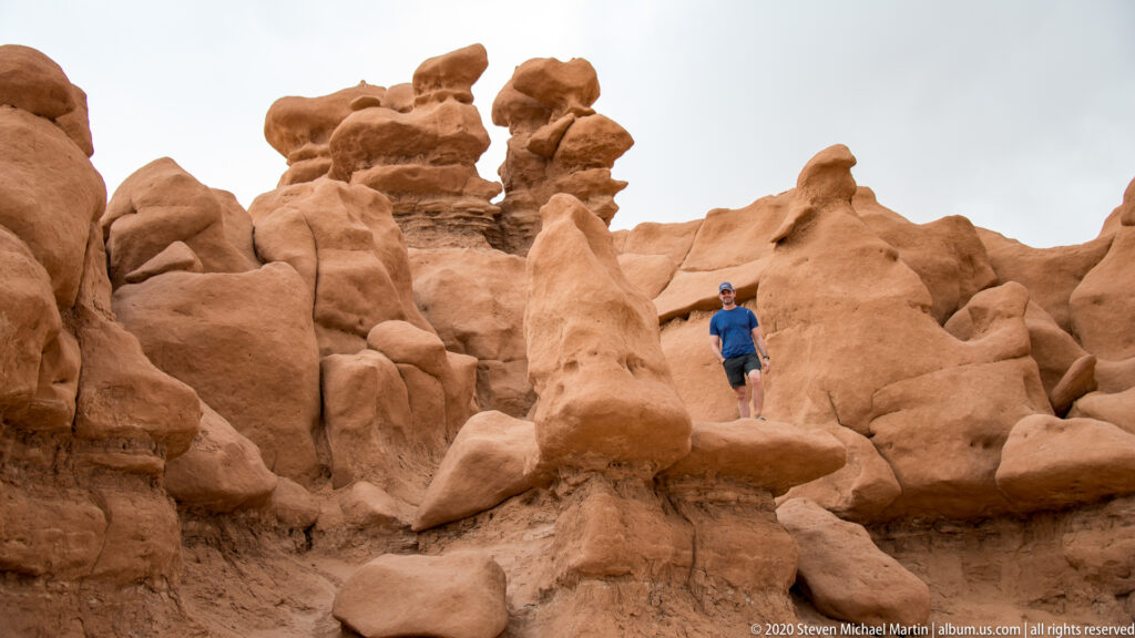 A visitor in a blue shirt stands on a narrow rise among tall rounded hoodoos.