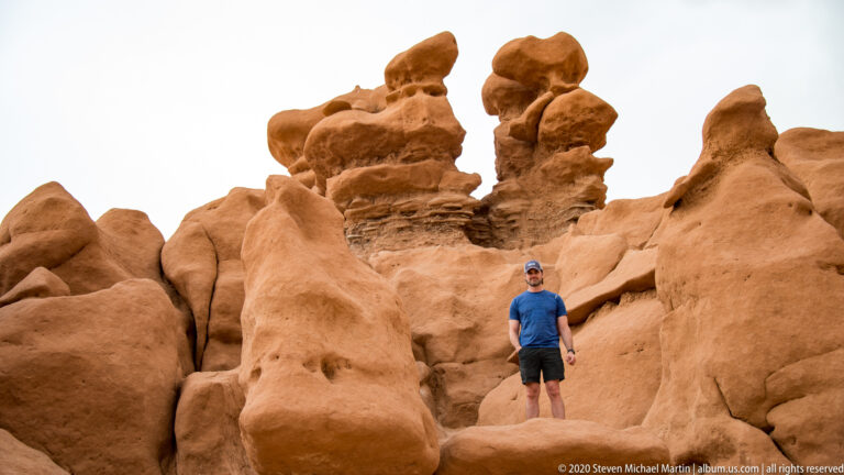 A visitor in blue and dark shorts stands between tall sandstone hoodoos.