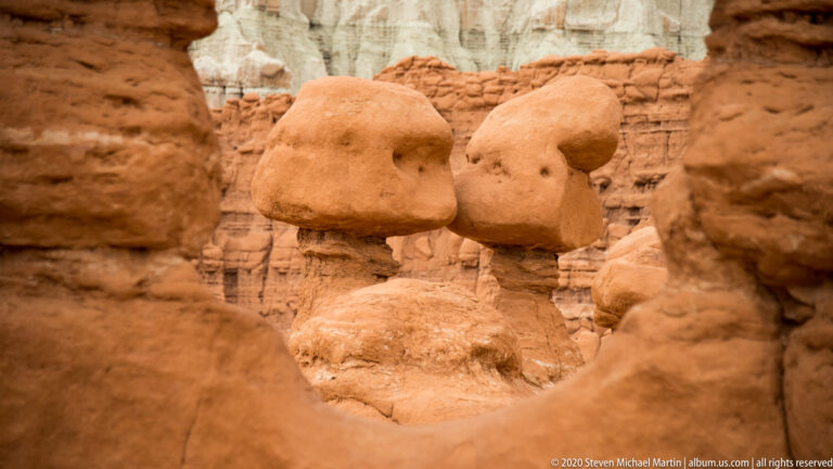 Two rounded hoodoos stand in a sandy passage before pale layered cliffs.