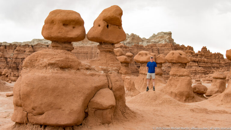 A visitor in blue stands among large rounded hoodoos with layered cliffs behind.