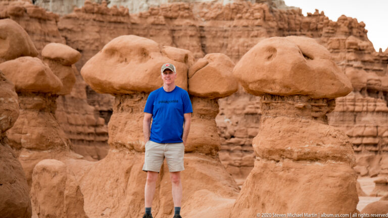 A visitor in blue and pale shorts stands among broad-capped sandstone hoodoos.