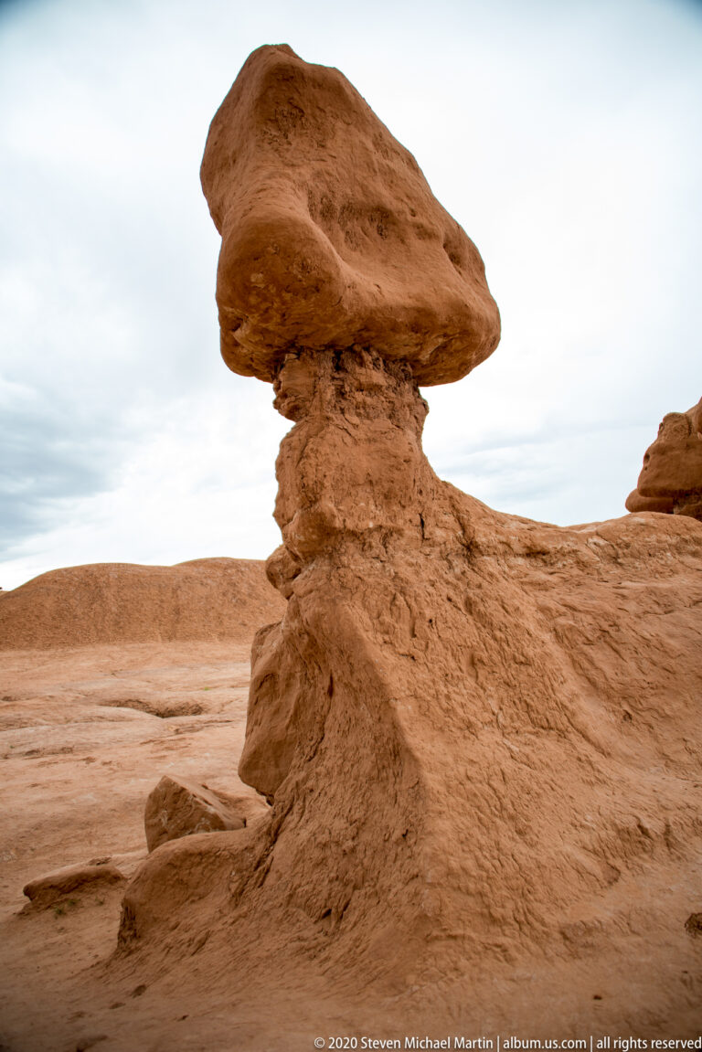 A broad capstone balances on a narrow pedestal above a smooth sandstone surface.