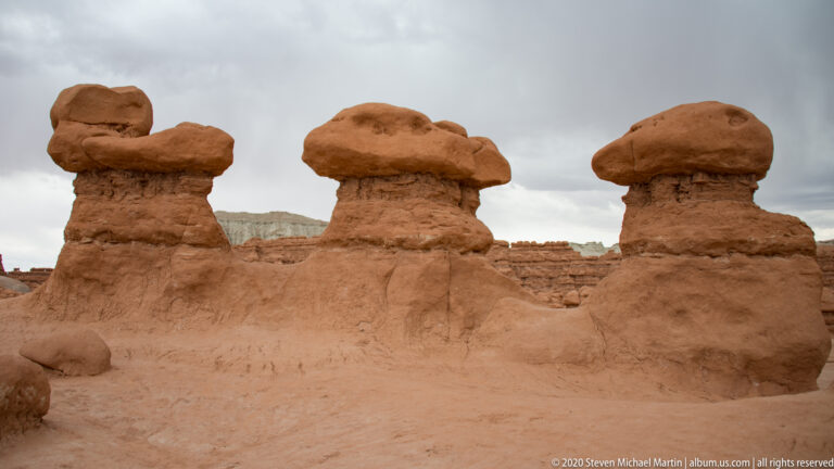 A short hoodoo and taller tower stand before a pale layered cliff and sandy ground.