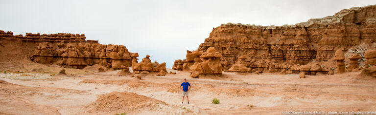 A visitor stands in a sandy basin beneath high sandstone walls and scattered hoodoos.