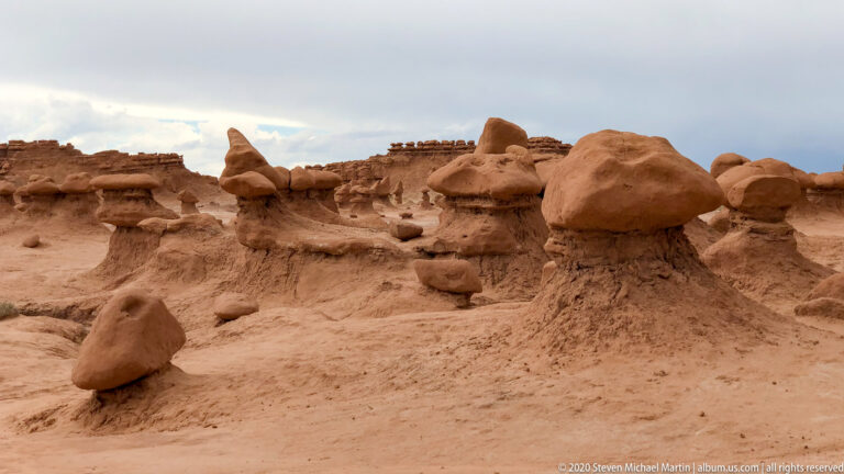 Low mushroom-shaped sandstone formations are scattered across an open pale basin below a layered cliff.