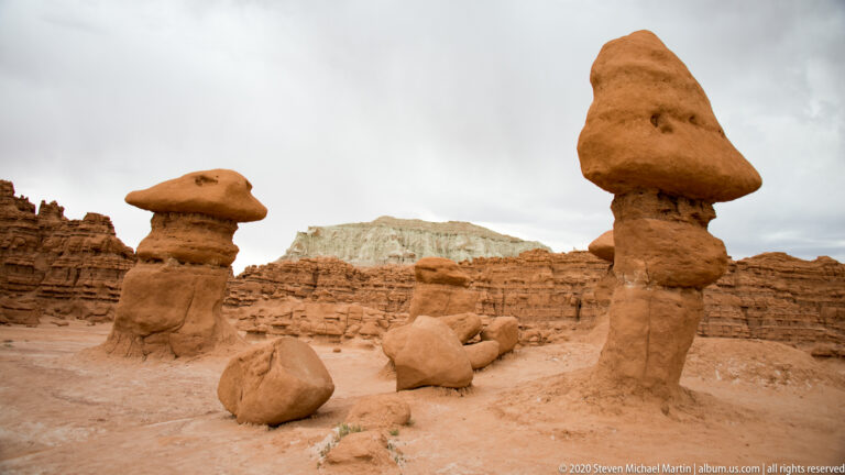 A short hoodoo and a taller tower stand before a pale layered cliff.