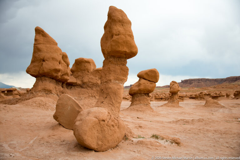 A tall foreground spire leads toward smaller capped hoodoos across a sandy basin.