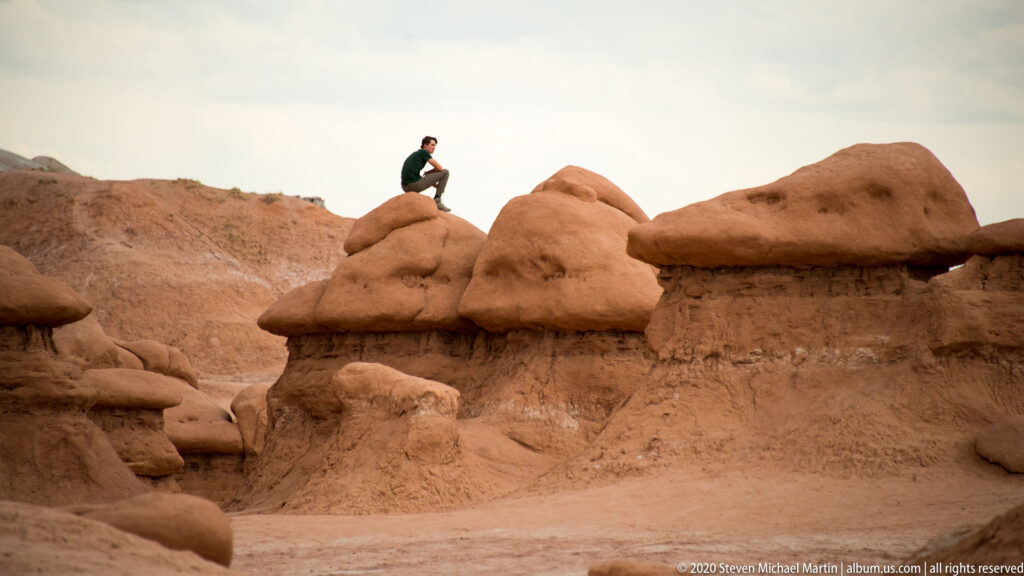 A visitor sits atop a rounded caprock above a sandy passage between hoodoos.