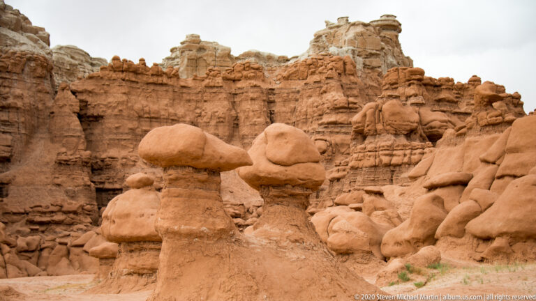 A broad-capped hoodoo stands before smaller pedestals and a banded sandstone wall.