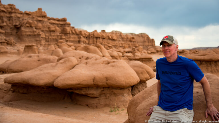 A visitor in blue stands beside a broad-capped hoodoo beneath layered cliffs.