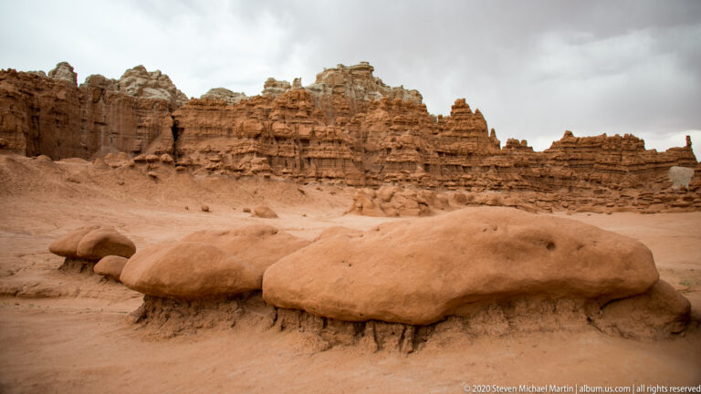 A low flattened rock mound lies below a long stratified cliff beneath a gray sky.