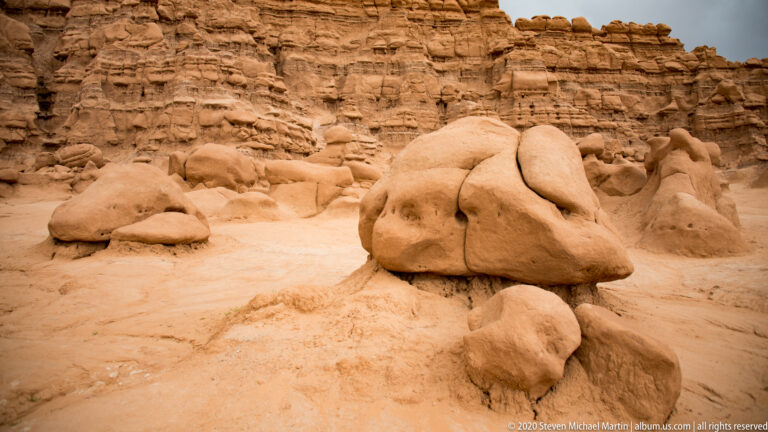A large rounded caprock rises above smaller boulders and pedestals on a sandy floor.