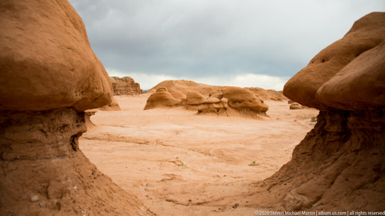 Large hoodoos frame a narrow sandy passage leading toward smaller rocks and cliffs.