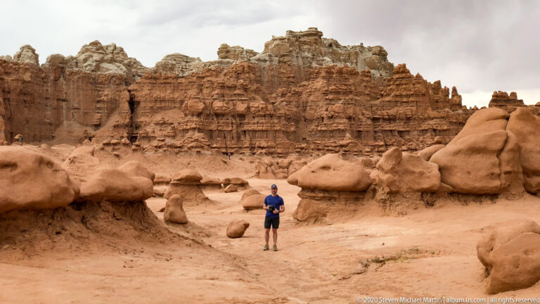 A visitor stands among low hoodoos in a sandy basin beneath a tall layered sandstone cliff.