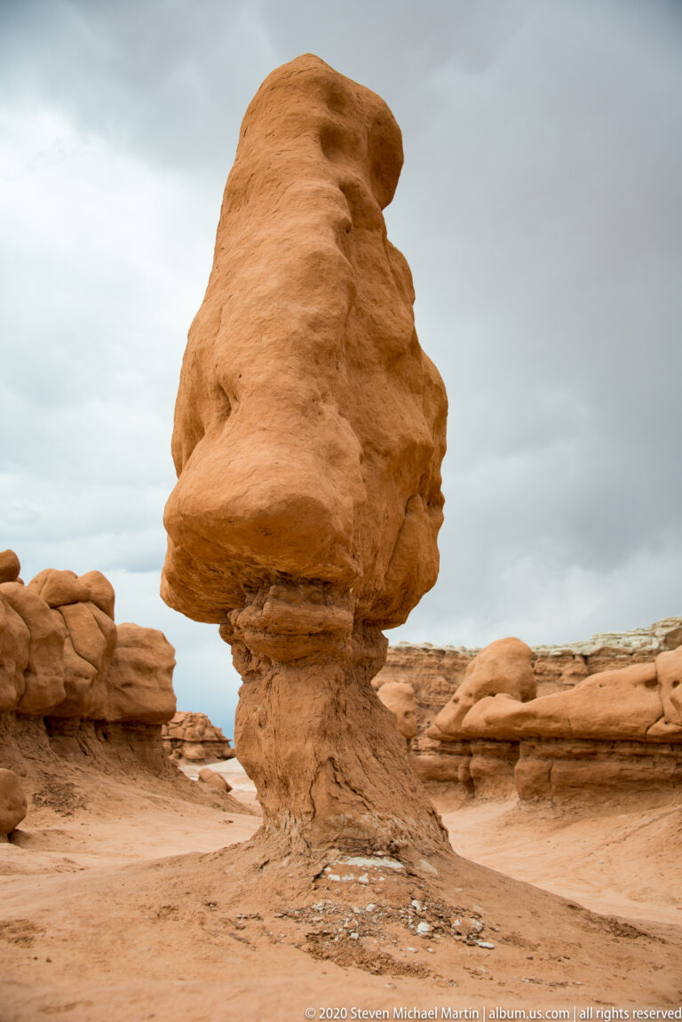 A massive uneven capstone overhangs a narrow pedestal on a smooth sandy floor.