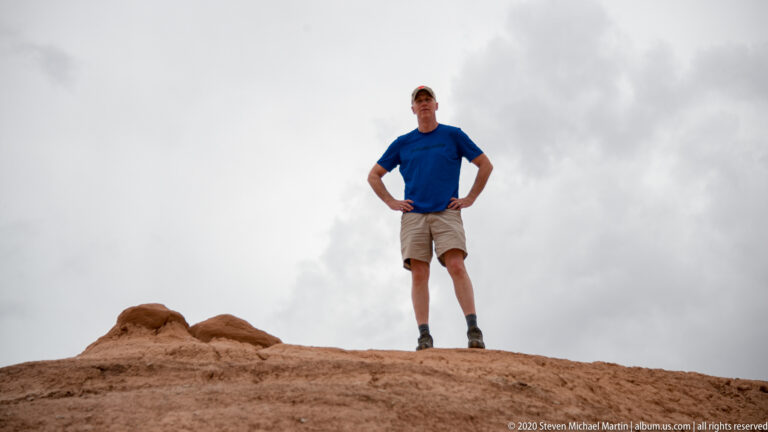 A hiker stands with hands on hips atop a rounded sandstone crest beneath a cloudy sky.