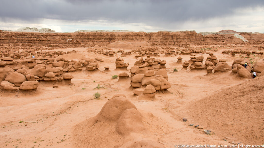 Visitors walk among rounded sandstone hoodoos across an open valley beneath a flat ridge.
