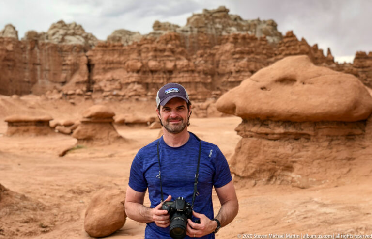 A visitor holds a camera in front of sandstone hoodoos and a banded cliff.