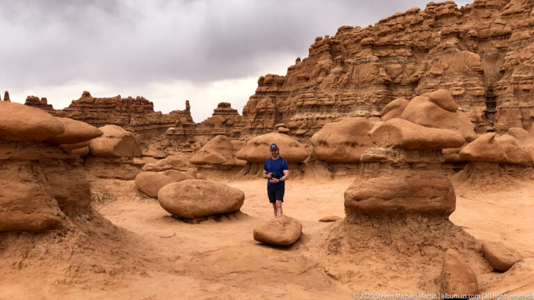 A visitor walks along a sandy route between large hoodoos beneath a layered cliff.