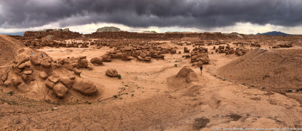 A vast field of sandstone hoodoos lies beneath dark storm clouds, with visitors visible in the distance.
