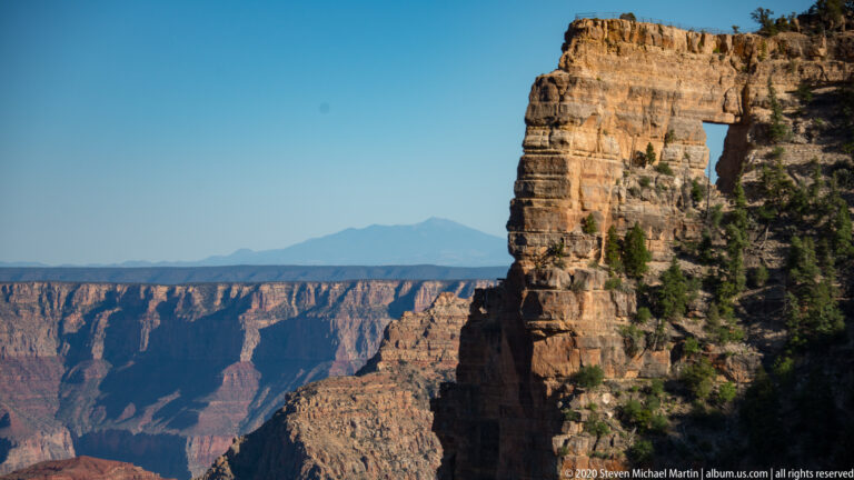 A large opening in a tan rock promontory frames distant canyon walls beneath a clear sky.
