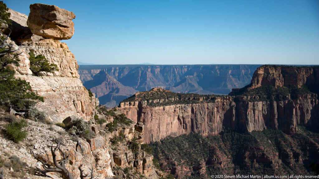 A pale foreground cliff borders a green-topped ridge and broad red canyon walls under a blue sky.