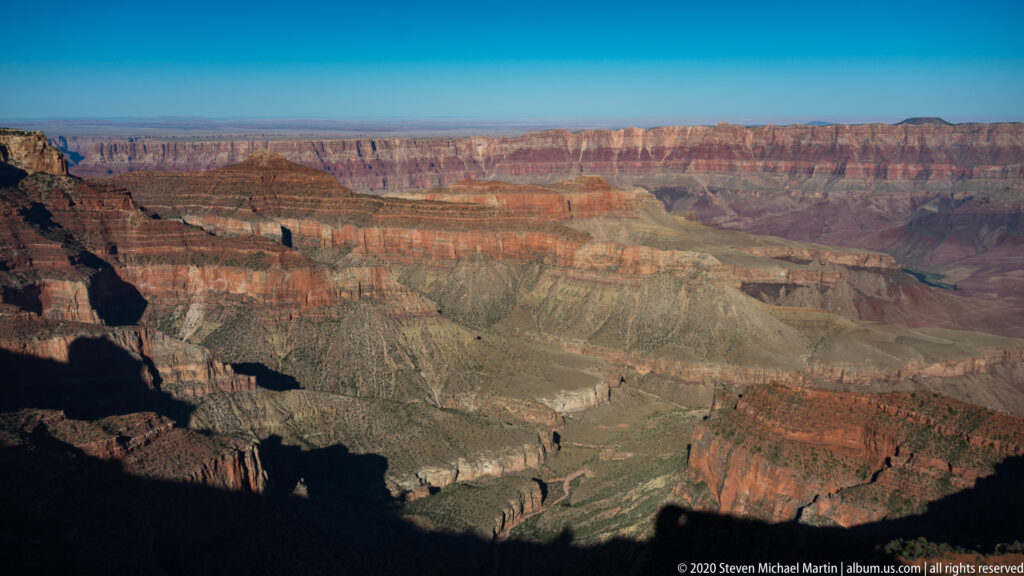 Layered red, ochre, gray, and green canyon walls recede toward a level distant rim.