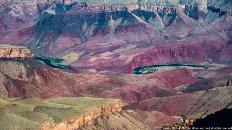 A narrow green stretch of the Colorado River winds between pink, purple, and red canyon slopes.