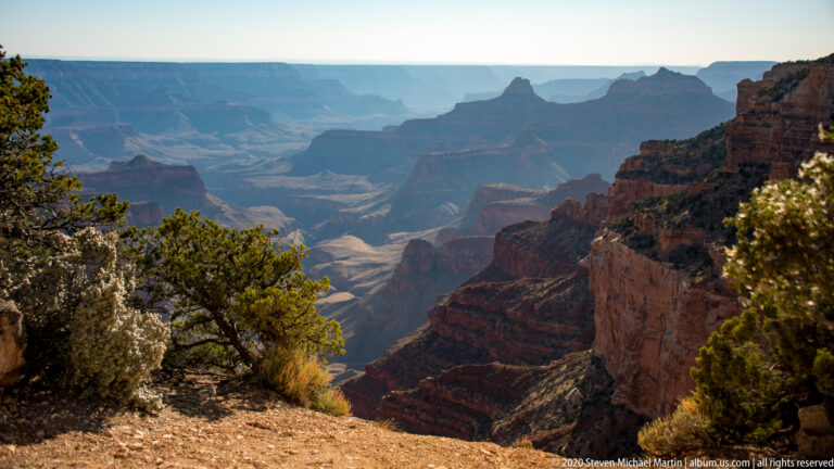 Rim shrubs frame hazy red canyon walls and distant mesas fading into blue atmospheric light.