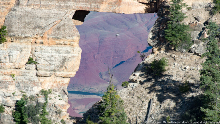 A small blue-green river section lies far below a pale rim ledge and scattered conifers.