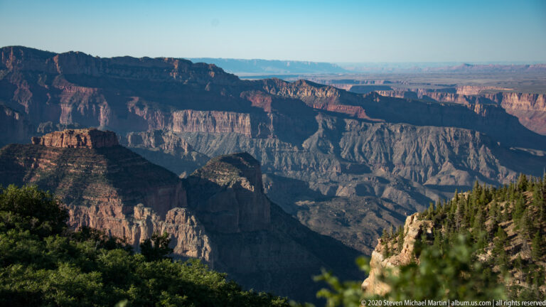 Dark rim trees frame sunlit red and gray canyon walls stretching toward a distant plateau.