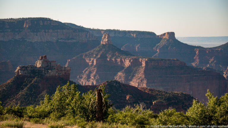 Two isolated rock spires rise above dark green ridges against distant canyon walls in warm light.