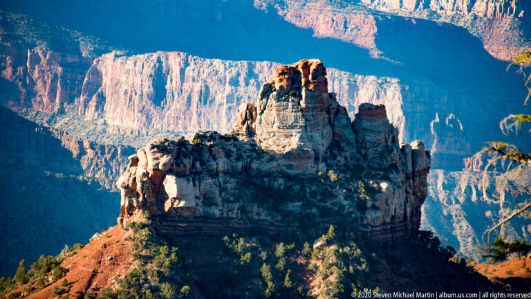 A pale, banded rock tower rises from a red slope against shadowed canyon walls.