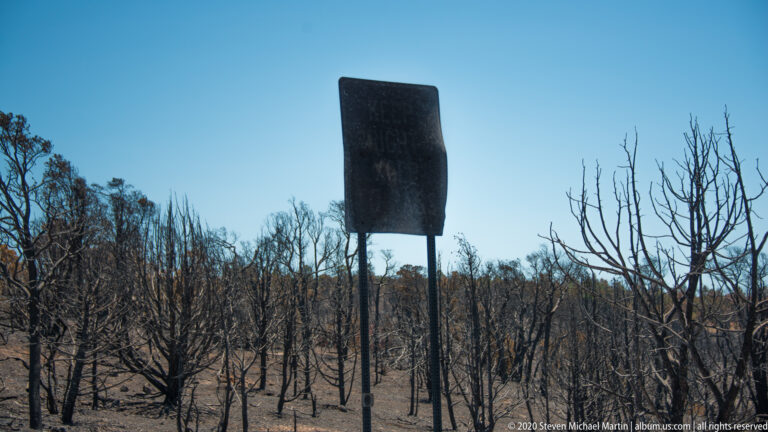 The back of a dark rectangular sign stands among charred trees on a burned slope.