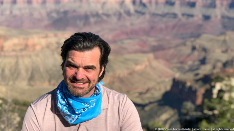 A man wearing a blue patterned neck gaiter poses with the blurred Grand Canyon behind him.