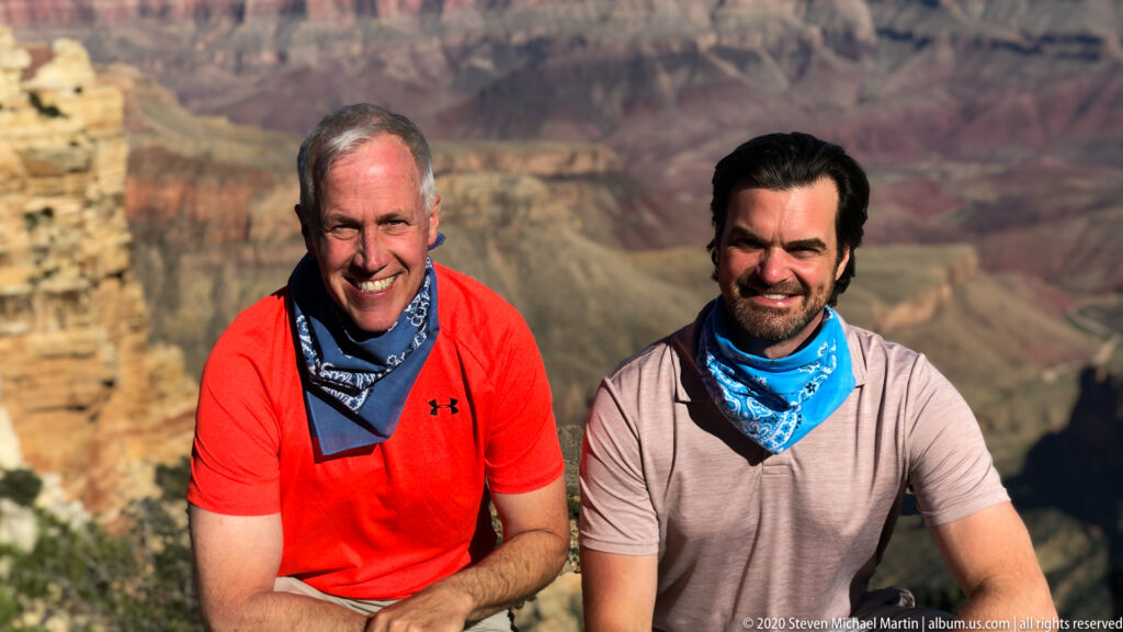 Two men pose together at the rim, with softly focused canyon cliffs behind them.