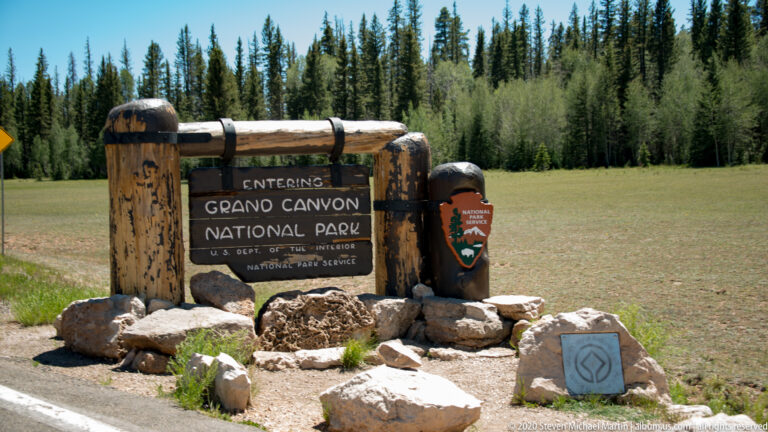 A timber sign reading “Entering Grand Canyon National Park” stands beside a meadow and conifers.