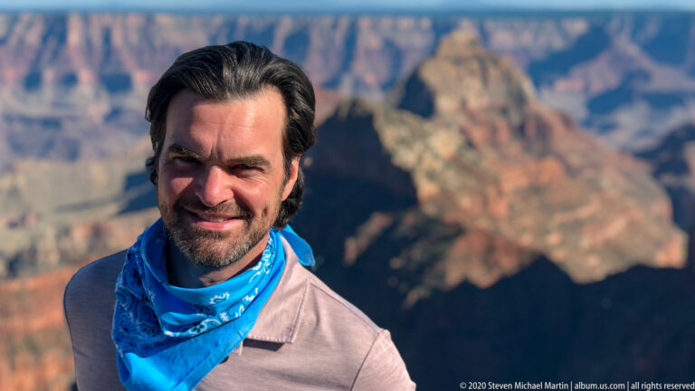 A smiling man in a blue neck gaiter poses against softly blurred canyon cliffs.