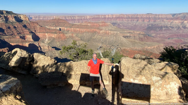 A visitor in a red shirt stands beside a stone rim barrier above the Grand Canyon.