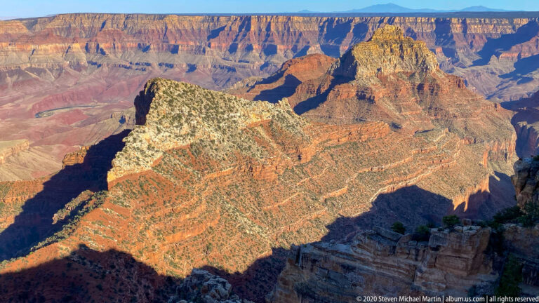 A pale-topped promontory projects into a deep canyon of orange, cream, and violet rock layers.