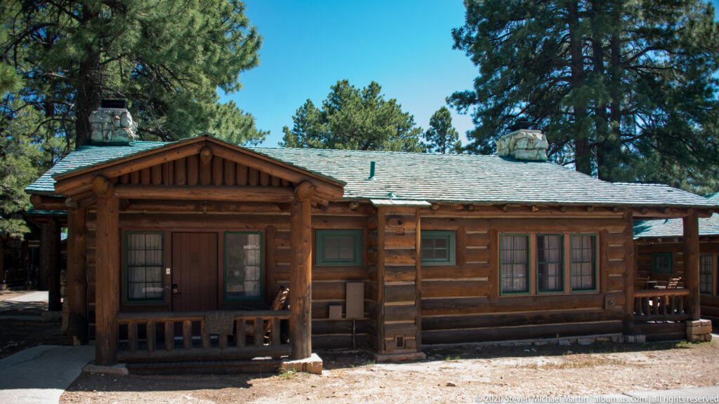 A low log building with a covered porch stands in a clearing among tall pines.