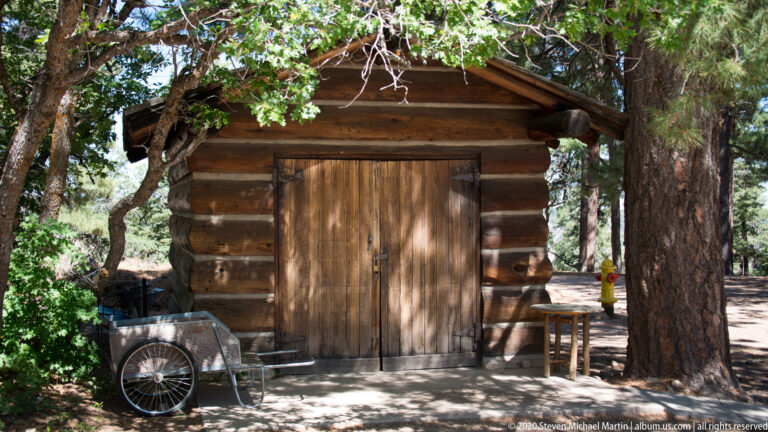 A small log outbuilding with closed double doors stands beneath trees beside a metal handcart.