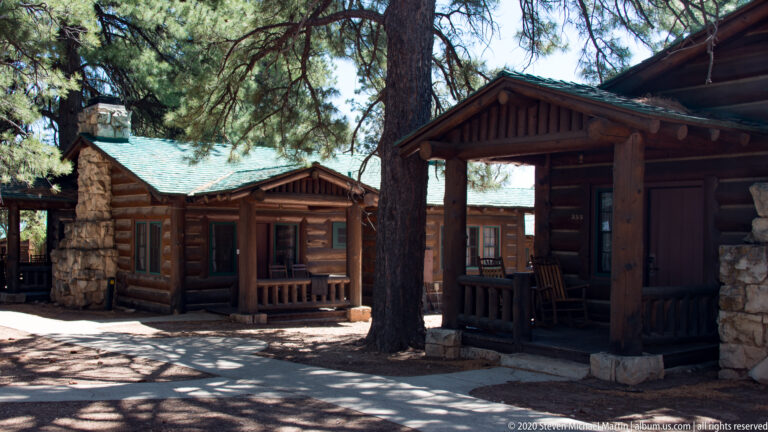 Log cabins with stone chimneys and covered porches stand in deep shade beneath mature pines.